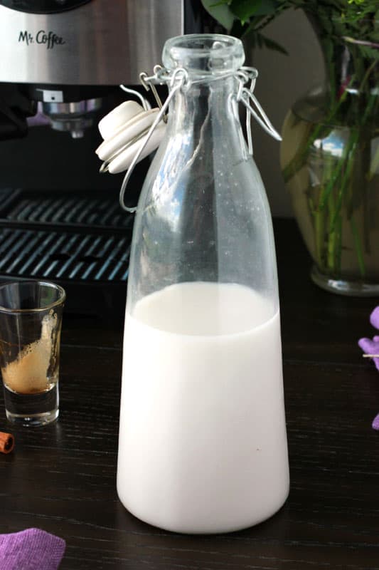 tall glass container that has an attached lit filled with homemade horchata. The container is sitting on top of a dark wooden table top with an espresso machine sitting in the back. Next to the machine is a glass shot glass that was once filled with espresso and shows the remaining foam. 