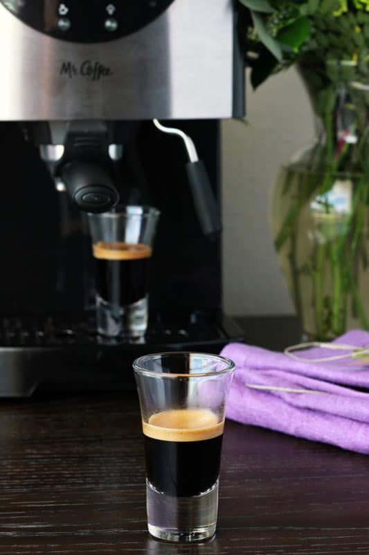 short shot glass sitting on a dark table top filled with espresso and topped with that classic foam. Another shot glass filled with espresso is sitting on top of the espresso machine in the background. A stack of purple napkins and a vase filled with florals are off to the side