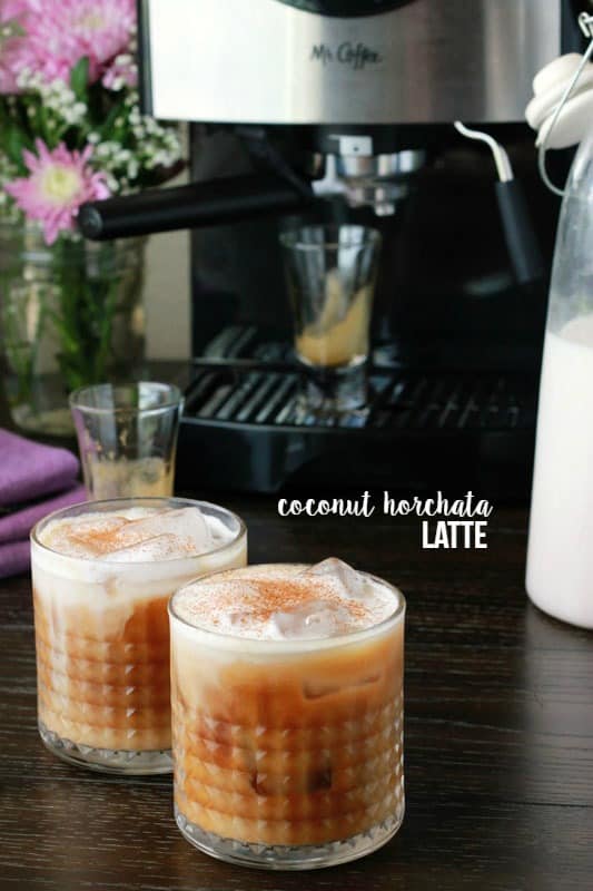 two short glasses filled with ice, espresso and topped with a foam. Cups are sitting on a wooden table top with an espresso machine in the background with two empty glasses of espresso. Text overlay reads "coconut horchata latte"