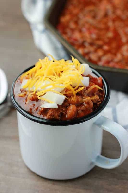 close up of a white camping tin mug filled with chili and topped with shredded cheese and diced onions