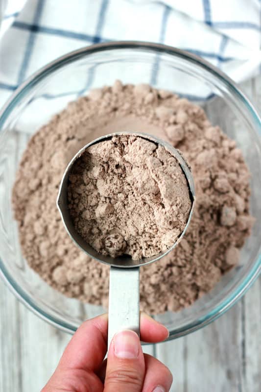 Measuring cup hovering over a glass mixing bowl that is filled with brownie baking mix