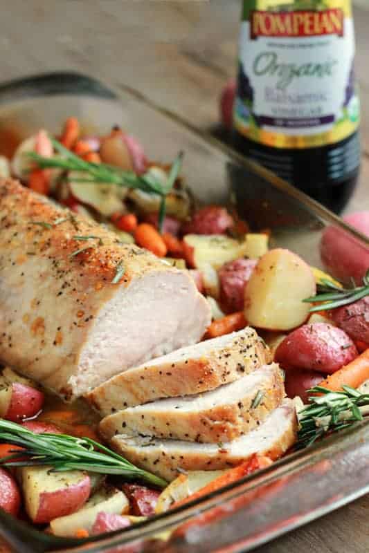 close up image of a pork tenderloin that has been cooked and sliced on top of a bed of fresh rosemary, red potatoes, and baby carrots.