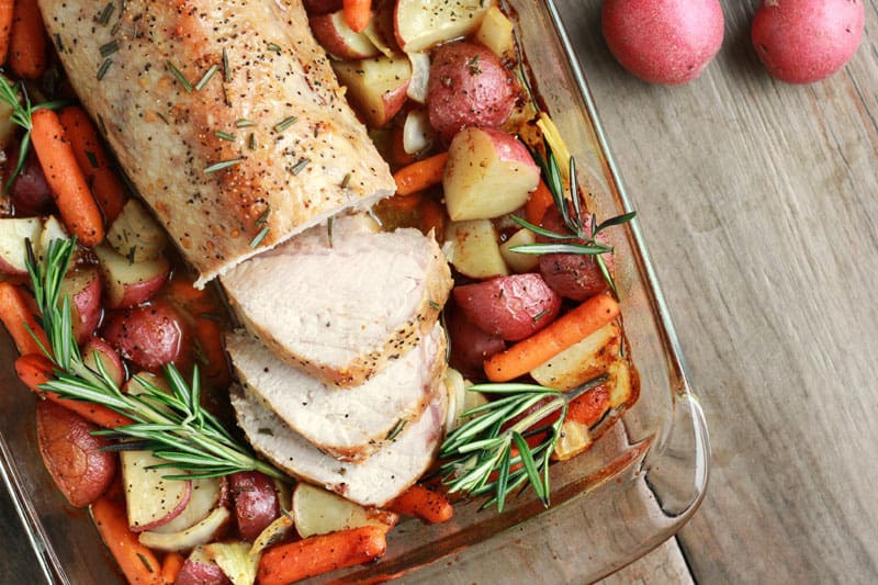 top down image of a 9x13 pan that is filled with cooked potatoes and carrots and topped with a cooked pork loin that has been sliced and fresh rosemary
