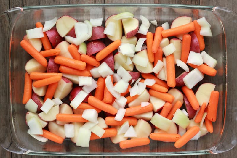 Uncooked chopped potatoes, baby carrots and onions sitting in a 9x13 dish on a light brownish grey table