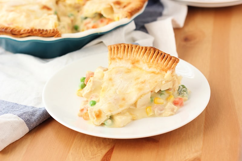 Slice of chicken pot pie sitting on a white plate with the rest of the pie off to the back left corner.
