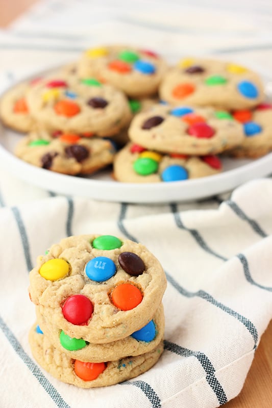 three mm cookies stacked on a white and blue striped napkin with a round white plate in the background stacked with more cookies