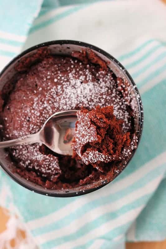 top down image that is close to the top of a mug that is filled with a chocolate cake that has been dusted with powdered sugar. A bite is being pulled out on a spoon