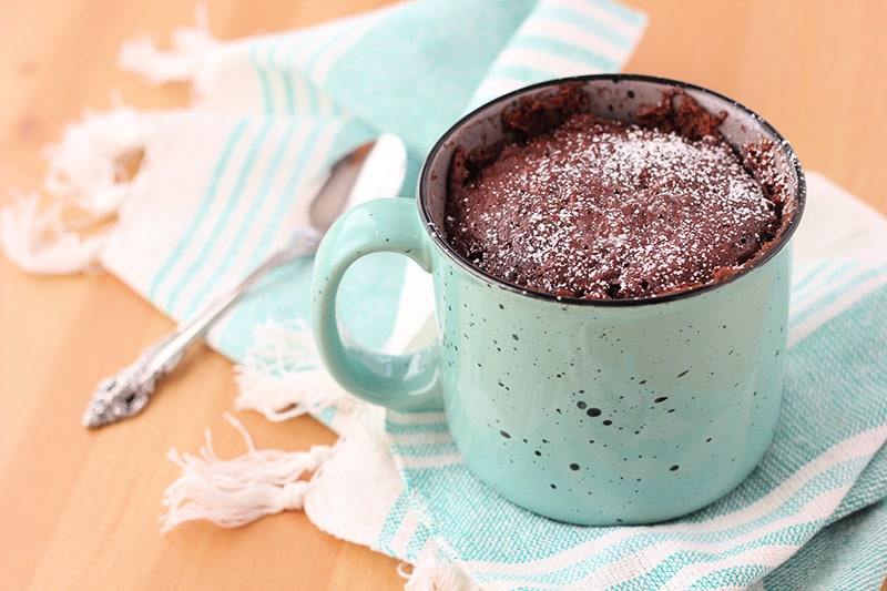 A blue speckled mug with a chocolate cake inside that has been dusted with powdered sugar. Mug is sitting on a light blue and white striped napkin on a wooden table top