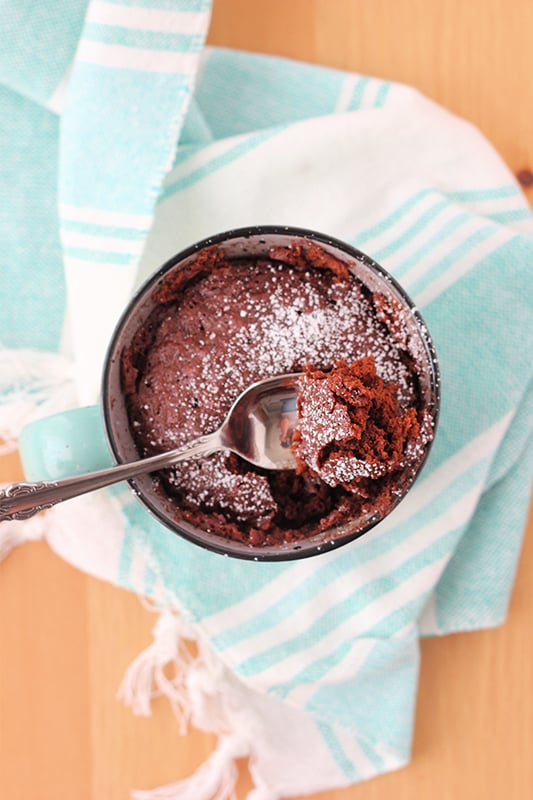 top down image showing a mug that is filled with a chocolate cake that has been dusted with sugar with a bite on a spoon. There is a light blue and white striped napkin under the mug and everything is sitting on a wooden table