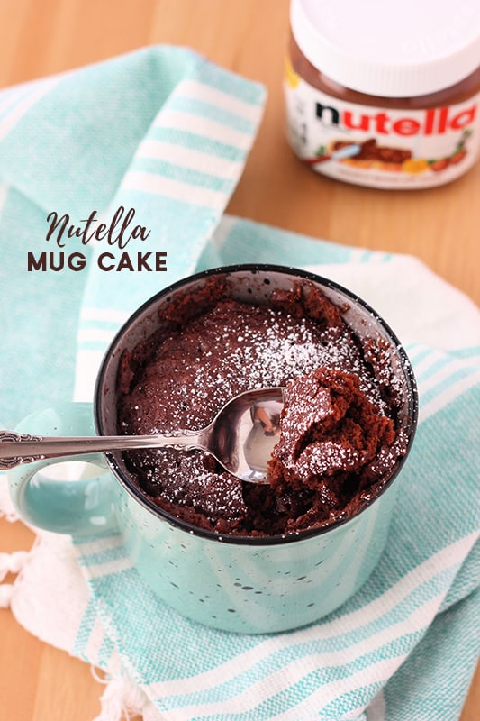 Chocolate cake inside a light blue speckled mug with a bite being pulled out on a spoon. The mug is sitting on a light blue and white striped napkin on a wooden table with a container of Nutella sitting in the back