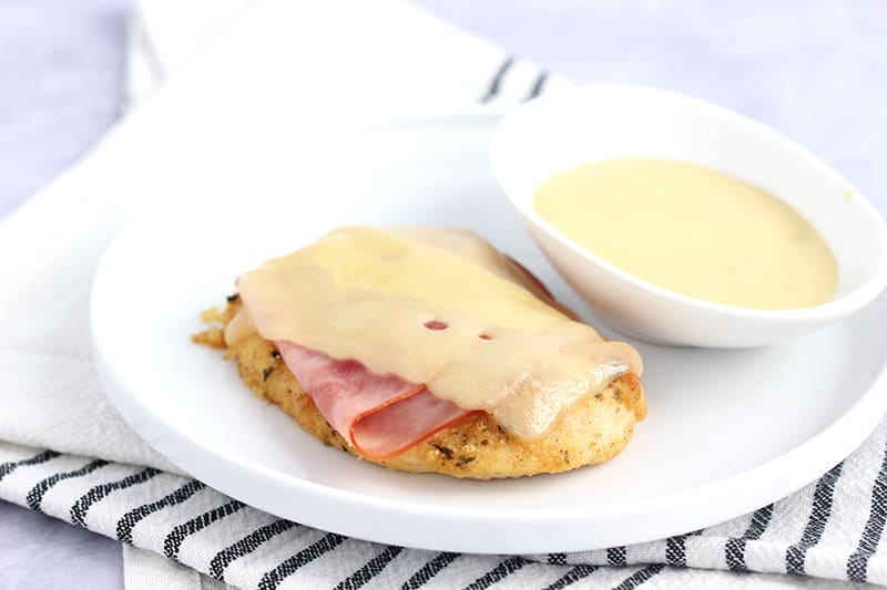 White plate with a slight raised edge topped with a piece of baked chicken that has a slice of ham and melted cheese directly on top. On the side is a white bowl filled with mustard dip