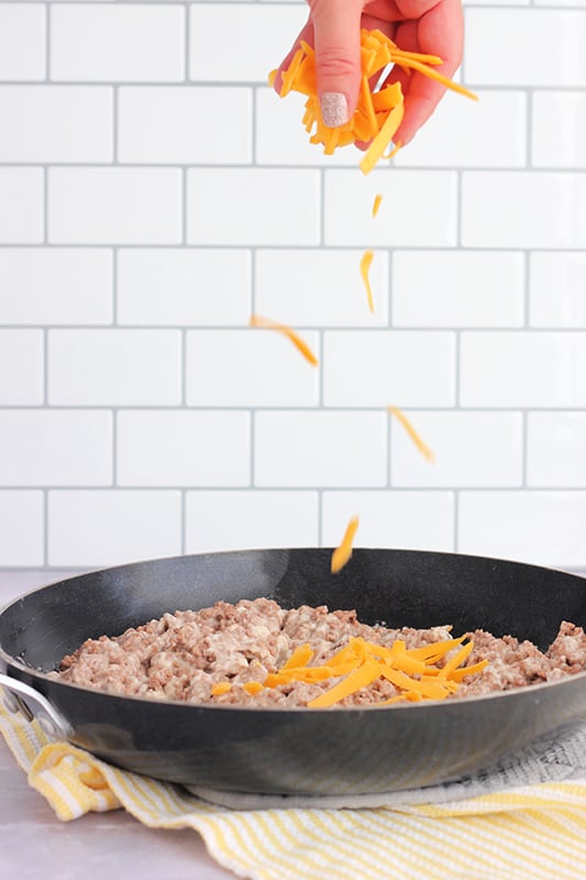 black skillet sitting on a white countertop with ground beef inside. A hand is sprinkling shredded cheese over the top