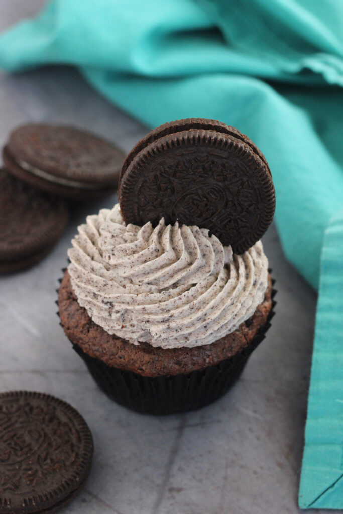 single oreo cupcake sitting on a grey background with a teal napkin and extra chocolate sandwich cookies sitting off to the side