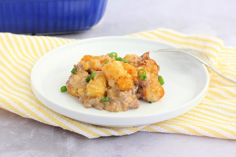 small white plate with a lifted edge sitting on a yellow and white striped napkin with a serving of tater tot casserole sitting on top. A fork is off to the side with a blue baking dish sitting in the background