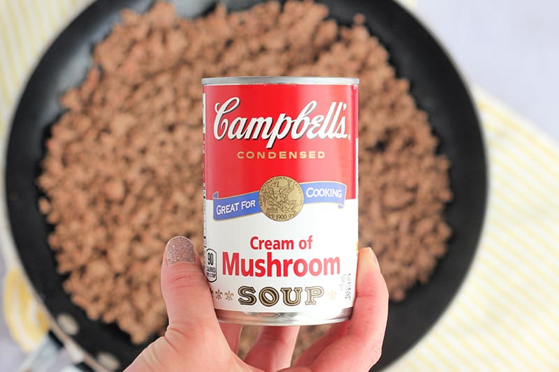 hand holding a can of Campbell's cream of mushroom soup over a black skillet that is filled with browned ground beef