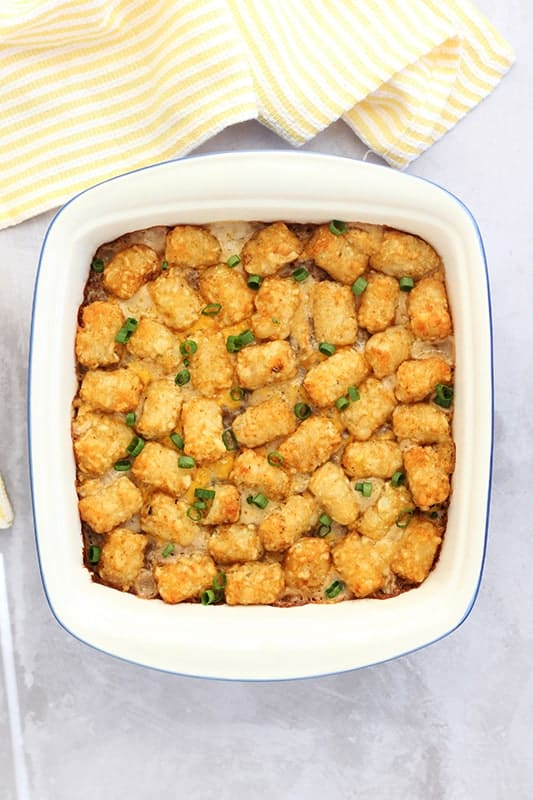 top down image of a 9 inch white baking dish that is filled with a casserole that is topped with crispy baked tater tots. A white and yellow striped napkin is off to the side of the white counter