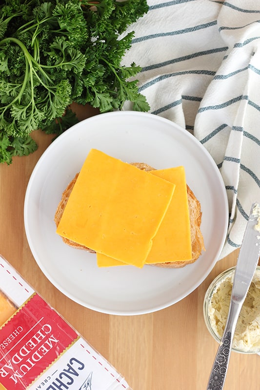 top down image showing two slices of cheese sitting on top of a slice of bread on a white round plate. The plate is sitting on a wooden table top that has fresh parsley, a stripe napkin, and a container of Cache valley medium cheddar slices off to the side of the plate. 