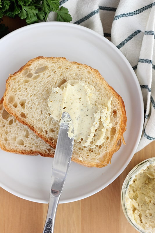 top down image showing a white plate with two slices of white bread being spread with a garlic butter using a butter knife