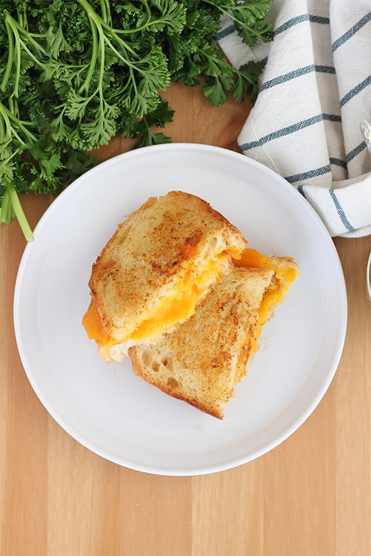 top down image showing a white round plate with a cheese sandwich that is sitting on a wooden table top with fresh parsley and a stripe napkin off to the side