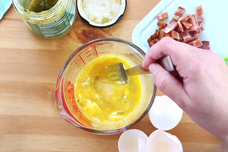 top down shot showing a hand whisking eggs in a glass measuring cup. Additional ingredients, like bacon and salsa, are sitting off to the side on a wooden table top