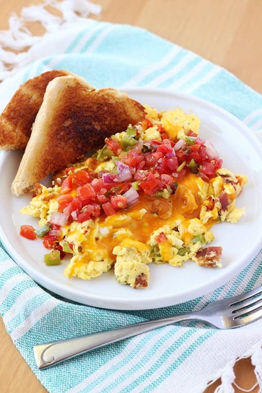 close up image of a small white round plate with a lifted edge topped with scrambled eggs that are filled with bacon and chopped peppers. On top of the eggs are melted cheese and pico and a side of triangle cut toasts off to the side