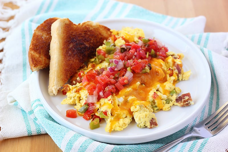 small round white plate that is topped with scrambled eggs that are loaded with vegetables and bacon. Topped with melted cheese and fresh pico with a side of toast