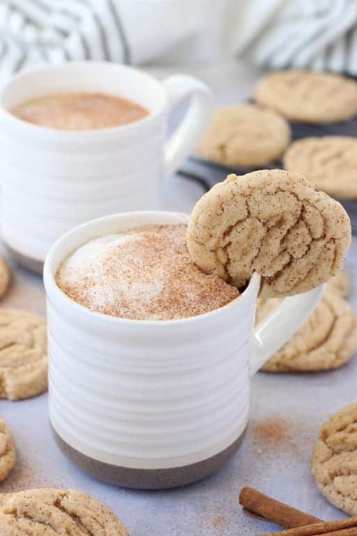 white mug with a dark tan bottom that is filled with coffee, frothed milk, and cinnamon sprinkle with a snickerdoodle cookie hanging off the edge.  A second cup of coffee off in the background with more cookies and cinnamon sticks on the table top