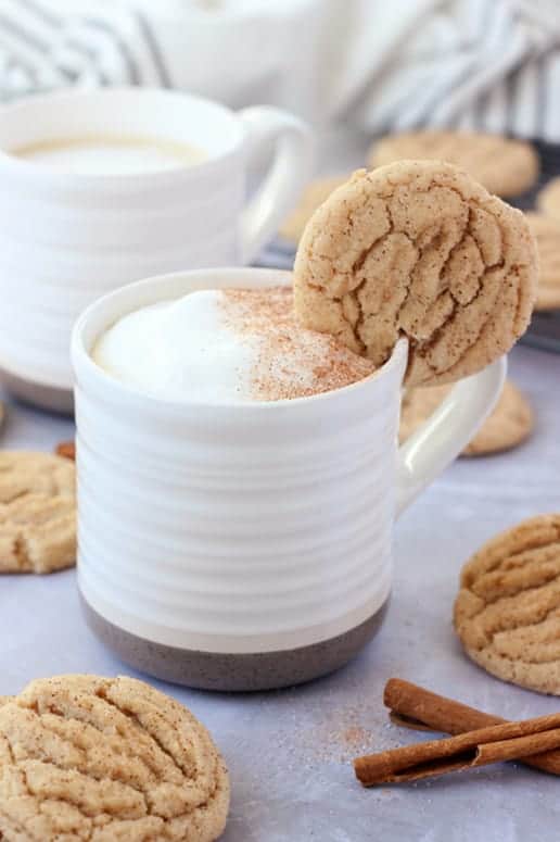 white mug with a dark tan bottom that is filled with coffee, frothed milk, and cinnamon sprinkle with a snickerdoodle cookie hanging off the edge.  A second cup of coffee off in the background with more cookies and cinnamon sticks on the table top