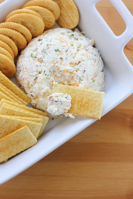 top down image showing a cheese dip in the shape of a ball in a white rectangle serving dish with crackers spread around the dip. One cracker sitting on top of the round with cheese spread onto the corner.