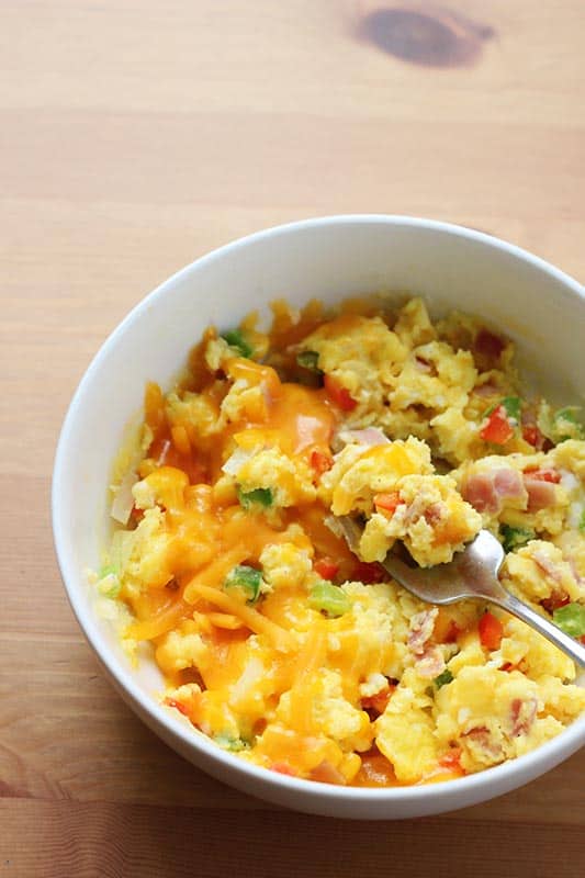 close up of a white bowl with fluffy scrambled eggs topped with peppers, ham, and cheese with a bite on the fork
