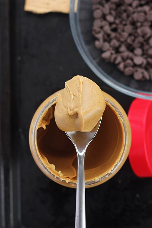 close up image of a jar of creamy peanut butter with a scoop sitting on a spoon balancing on top of the open container