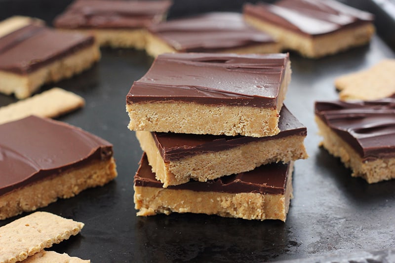 three no bake peanut butter chocolate bars stacked on top of each other on a black baking tray with more bars scatered around