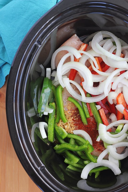black slow cooker insert filled with fajita ingredients like chicken breast, seasoning, and chopped peppers. 