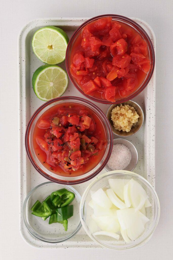 Top down image showing two small bowls of tomatoes in suace, a cut lime, chopped jalapeno, onions, garlic, and salt