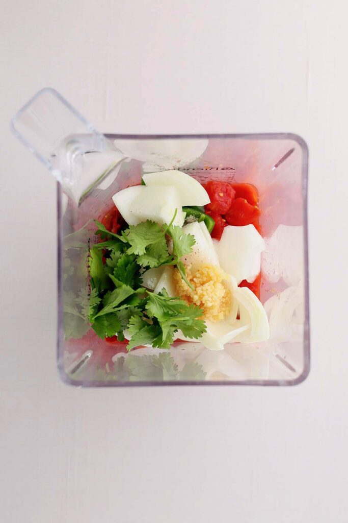 Top down image showing the insider of a blender with tomatoes, chopped onion, jalepeno, cilantro, and garlic