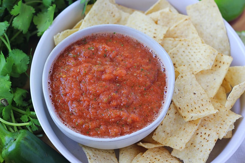 up close image showing a small white round bowl filled with salsa sitting in a larger bowl with tortilla chips surrounding the small bowl. Off to the side is a fresh jalapeno and additional cilantro