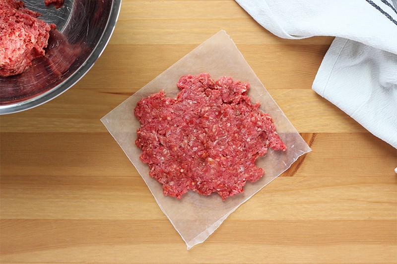 Ground beef shaped into a patty sitting on a wax paper square on top of a wooden table top with a silver bowl of more ground beef off to the side