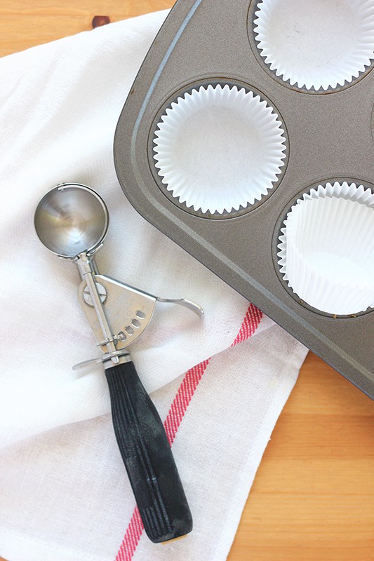 cupcake pan filled with paper liners sitting on a wooden table with a cookie scoop off to the side