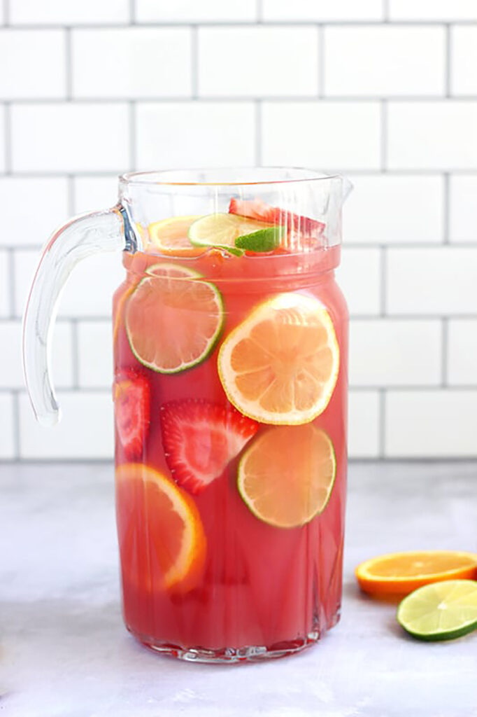 glass pitcher filled with a light red punch along with fresh fruit slices including strawberries, oranges, and limes
