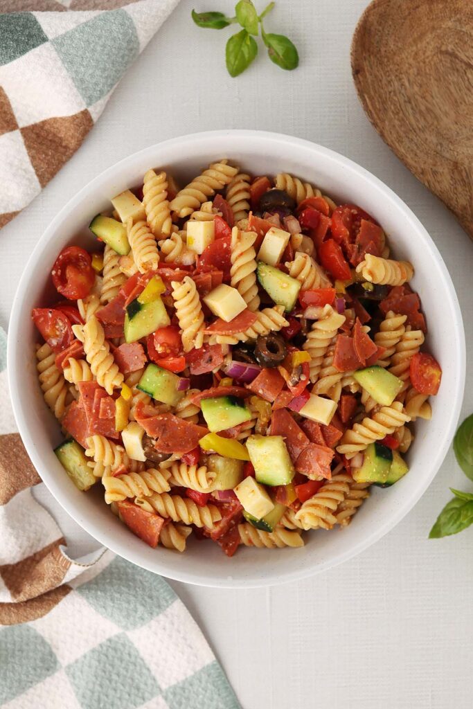 Top down section showing pasta salad in a white dish filled with spiral pasta and chopped veggies. A colored napkin, fresh basil, and a wooden spoon off in the back