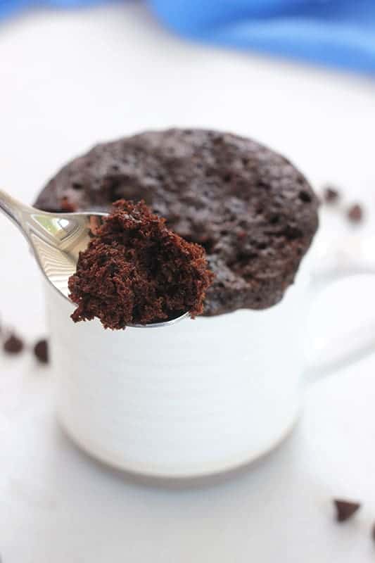 white mug with ridges with a tan bottom filled with a chocolate cake that is slightly overflowing with chocolate chips around the mug on a white table top with a bite on a spoon in front