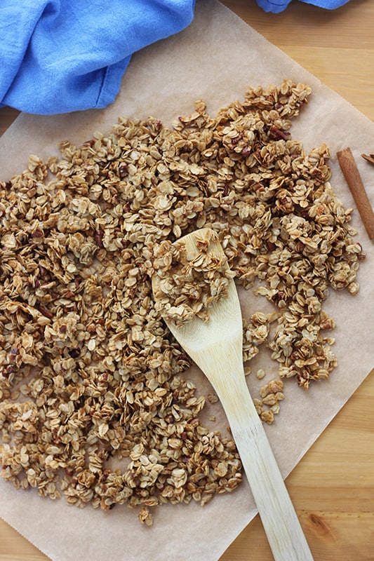 Top down image showing a parchment paper with a spread of homemade granola. A scoop is on a wooden spoon. There is a blue napkin and a cinnamon stick off to the side