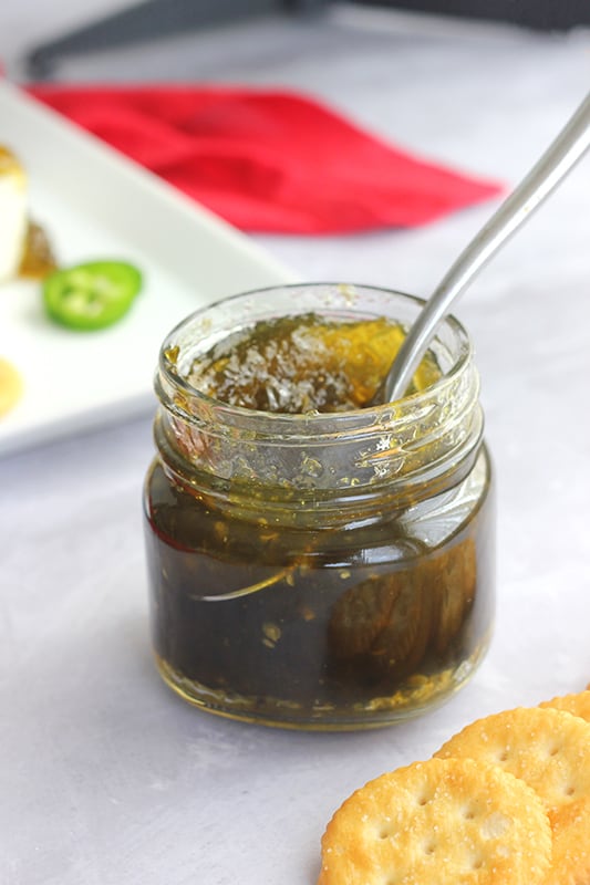 Jalapeno Jelly Recipe in a mason jar with a spoon inside the jar. Crackers, a white rectangle plate, and a red napkin are off the the sides.