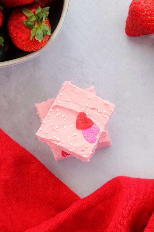 top down image showing two pieces of pink fudge that are cut into squares stacked on top of one another on a light gray table top with a bowl of fresh strawberries and a red napkin off to the side