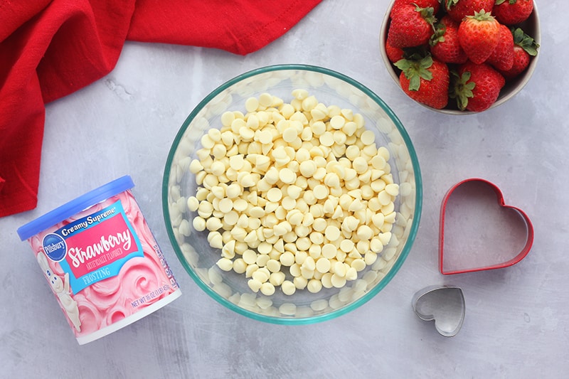 top down image showing a giant glass bowl filled with white chocolate chips, a can of Pillsbury strawberry frosting, a bowl of fresh strawberries, two heart cookie cutters and a red napkin on a light gray counter