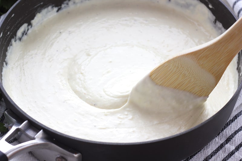 white alfredo in a black skillet being stirred by a wooden spoon