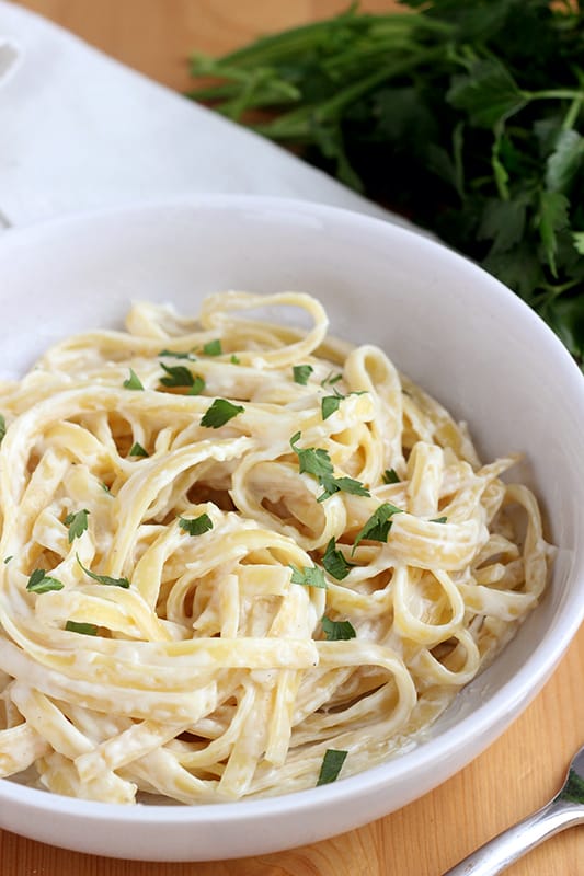 close up image of a bowl filled with cooked fettuccine noodles that are topped with a white creamy cheese sauce and fresh parsley pieces