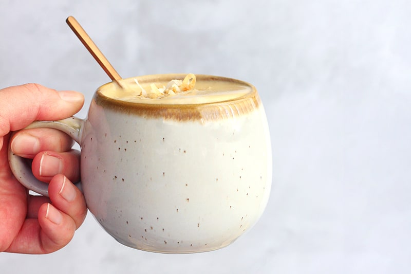 hand holding an off white speckled mug that is filled with a latte topped with toasted coconut pieces