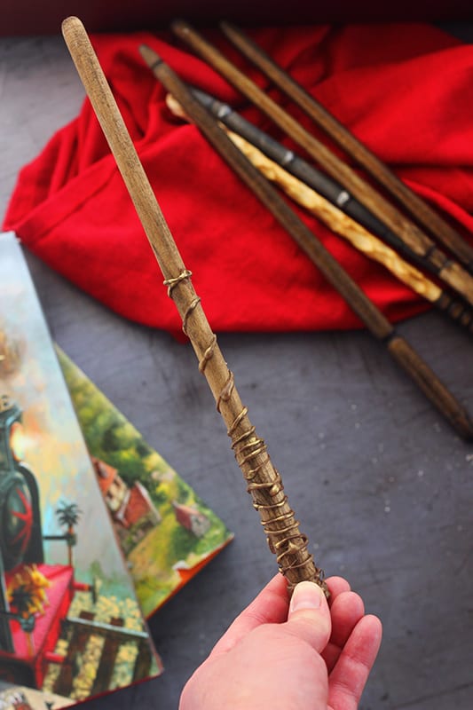 top down image showing a hand holding a wooden wand over top of a red napkin with more wands and the first two illustrated harry potter books on a dark grey table top