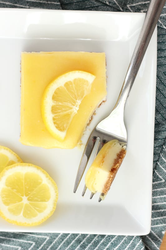 close up of a cheesecake bar topped with a lemon curd and slice of lemon on a white rectangle plate with a bite sitting on a fork. The plate is sitting on a blue chevron napkin